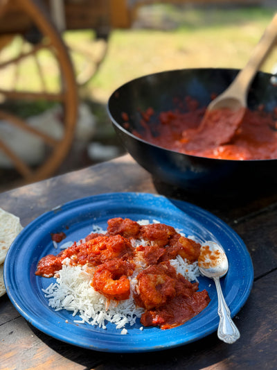 A blue plate of shrimp in a red-orange sauce over rice, on a picnic table next to a cast iron pot with more sauce and some flour tortillas.