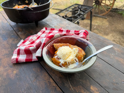 Cowboy Kent Rollins' Peach Cobbler in a silver bowl on a table with a red gingham napkin and cast iron dutch oven in the background.
