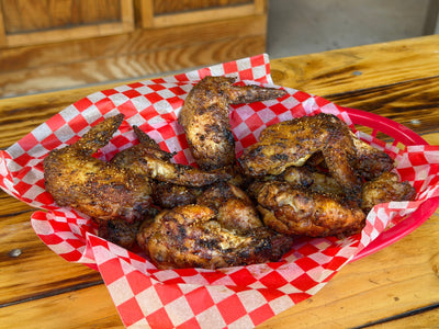 Photo is a basket of chicken wings that were prepared with apple cider vinegar mop sauce and a blend of seasonings.  The basket has wings, red checkered paper and is sitting on a wooden table.