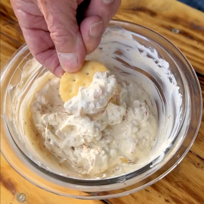 Image is a bowl of french onion dip and a hand holding a cracker that has been dipped