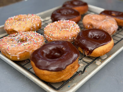 Cowboy Kent Rollins' Amish Style Donuts on a wire rack. Classic Glazed Donuts, donuts with sprinkles, and chocolate iced donuts.