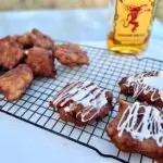 Kent Rollins Apple Fritters sit on a wire rack, three of them with icing. In the background is a bottle of Fireball Whiskey, used for icing.