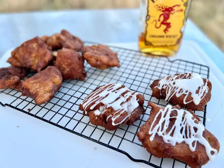 Kent Rollins Apple Fritters sit on a wire rack, three of them with icing. In the background is a bottle of Fireball Whiskey, used for icing.