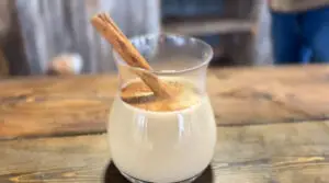 On a wooden table rests a small glass, halfway full of creamy egg nog. A dusting of cinnamon graces the top, and is garnished with a cinnamon stick for stirring.