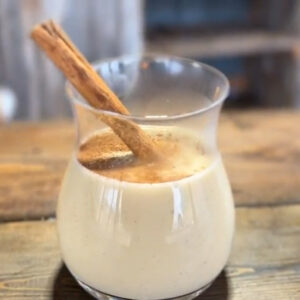 On a wooden table rests a small glass, halfway full of creamy egg nog. A dusting of cinnamon graces the top, and is garnished with a cinnamon stick for stirring.