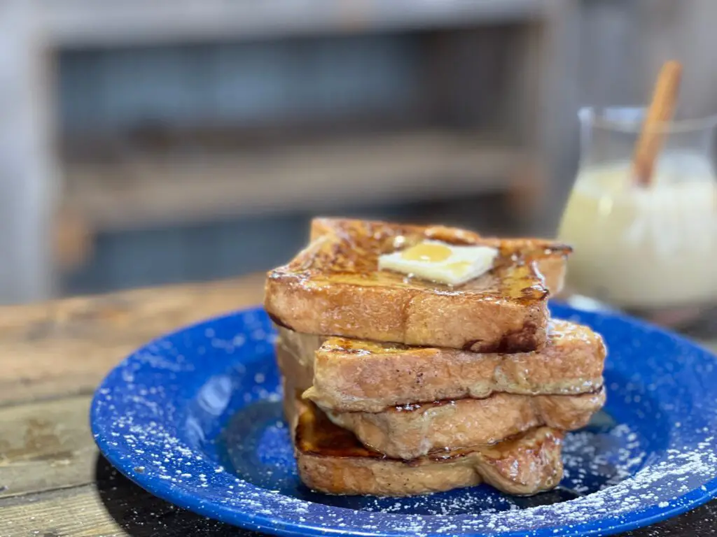 Four thick slices of egg nog french toast are stacked crookedly on a blue enamel plate. A thick pat of butter rests on top, and maple syrup cascades down the edges of the bread, pooling at the bottom.