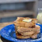 Four thick slices of egg nog french toast are stacked crookedly on a blue enamel plate. A thick pat of butter rests on top, and maple syrup cascades down the edges of the bread, pooling at the bottom.