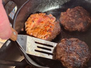 Three browned sausage patties cooking in a well-seasoned black cast iron skillet, a thin metal spatula is in the act of flipping one of the sausage patties. Steam can be seen rising from the skillet.