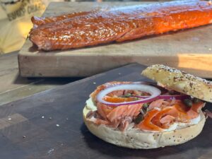 A sesame seed bagel rests on a mesquite cutting board, covered in smoked salmon, cream cheese, red onion, and capers. In the background rests a glazed, smoked salmon filet.