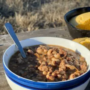 A white bowl with blue trim around the edge sits on a natural-oak colored picnic table. A bowl of black eyed pea soup, framed in steam, sits with a spoon resting on it's side. 