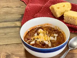 A white and blue bowl containing rich, hearty venison chili, garnished with shredded cheddar cheese and diced white onion. On a picnic table with a red tea towel and two slices of cornbread.