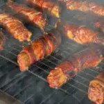 Amid a smoky atmosphere, 7 meat and cheese filled manicotti wrapped in bacon, aka shotgun shells, are crispy and delicious looking while sitting on a wire rack inside the Roughneck smoker by Kent Rollins