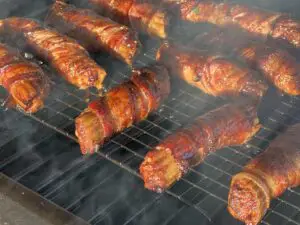 Amid a smoky atmosphere, 7 meat and cheese filled manicotti wrapped in bacon, aka shotgun shells, are crispy and delicious looking while sitting on a wire rack inside the Roughneck smoker by Kent Rollins