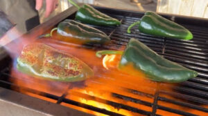 This photo shows five poblano peppers laying out on a grill, being blistered with a torch. One pepper is engulfed in flames and the skin is caramelizing and blistering.