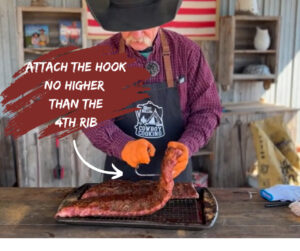 Photograph shows Cowboy Kent Rollins demonstrating how to put a metal hook through a rack of ribs by inserting it no lower than the 4th bone. 