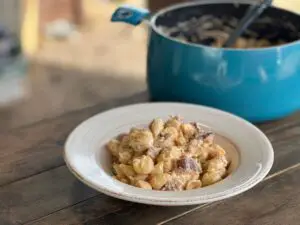 In the background, a blue enamel stock pot holds creamy, delicious smoked mac and cheese. In the foreground of the photo, a white plate holds smoked mac and cheese with smoked breakfast sausage. It is a miracle the pups didn't get to it before we got these photos taken.