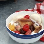 A white bowl with a blue stripe contains a banana split cobbler with melted ice cream all around the edges.