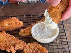 Four crispy golden fried fish rest on a wire rack, while the fifth is dipped in a thick homemade tartar sauce.