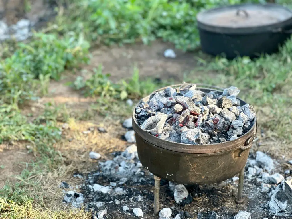 A Dutch Oven rests on a metal trivet. White hot coals on the lid and around the edge of the trivet are the heat source for the Dutch oven.
