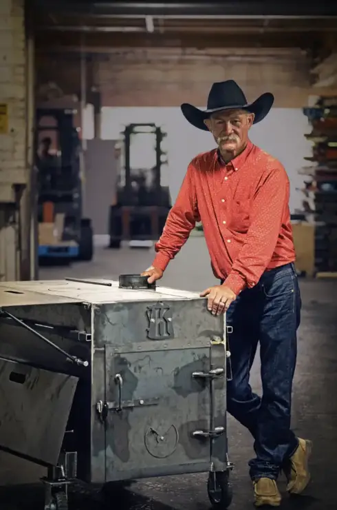 Man in a cowboy hat standing by a metal smoker.