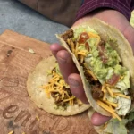 A person holding a taco with cheese and guacamole.