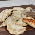 A wooden cutting board topped with dumplings and sauce.