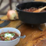 A bowl of chili next to an open cast fire pit.