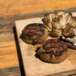 A wooden cutting board with some meat and an artichoke.