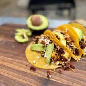 Cowboy Kent Rollins Bacon and Chorizo Breakfast Tacos, displayed on a wooden Mesquite cutting board. Corn tortillas, bacon, shredded cheese, queso fresco, and guacamole