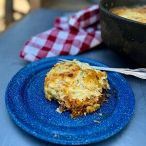 A blue enamel plate with cottage pie. In the background is a red checked cheesecloth and a Dutch oven. Cowboy Kent Rollins' Cottage or Shepherd's Pie