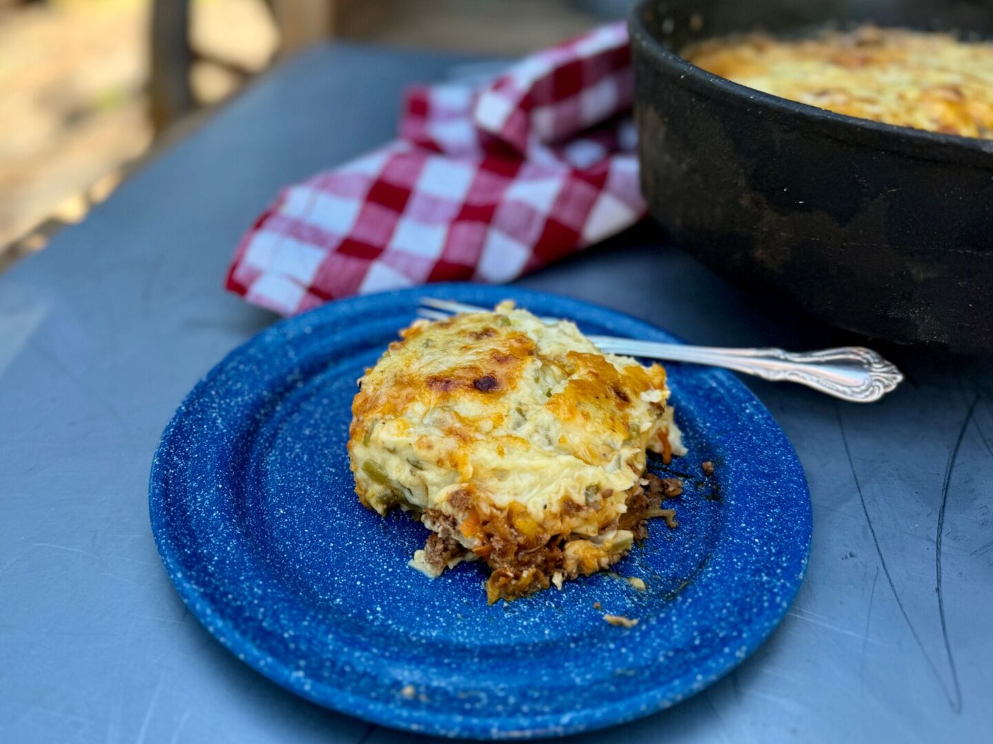 A blue enamel plate with cottage pie. In the background is a red checked cheesecloth and a Dutch oven. Cowboy Kent Rollins' Cottage or Shepherd's Pie