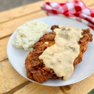 Cowboy Kent Rollins' Golden Crispy Chicken Fried Chicken with homemade gravy on a white plate, mashed potatoes on the side, with a red and white checked napkin in the background, on a picnic table.