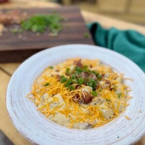 A bowl of loaded baked potato soup with heavy cream, cheddar cheese, bacon, and smoked sausage in a white bowl sits on a picnic table next to a green towel. In the background, green onions and shredded cheddar cheese sit on a dark cutting board.