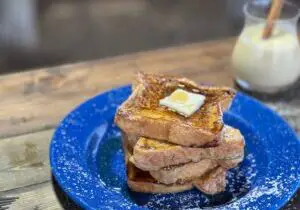 On a blue enamel plate, four thick slices of egg nog French toast are crookedly stacked, with a large pat of butter on top, and maple syrup cascading down the sides, forming a pool at the bottom.