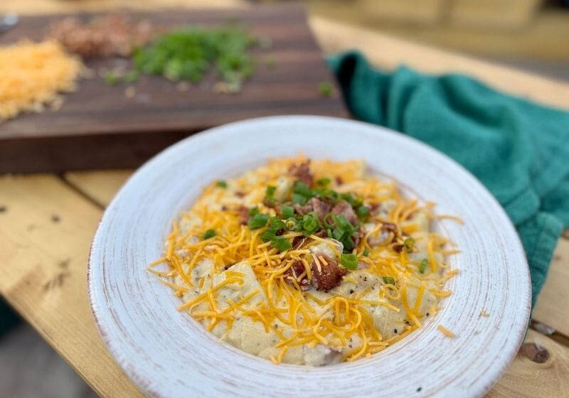 A bowl of loaded baked potato soup with heavy cream, cheddar cheese, bacon, and smoked sausage in a white bowl sits on a picnic table next to a green towel. In the background, green onions and shredded cheddar cheese sit on a dark cutting board.