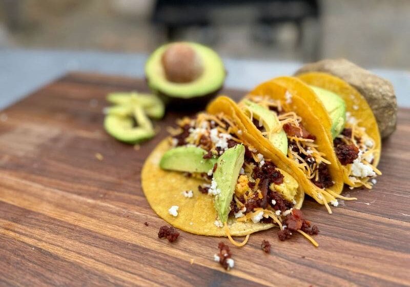 Cowboy Kent Rollins Bacon and Chorizo Breakfast Tacos, displayed on a wooden Mesquite cutting board. Corn tortillas, bacon, shredded cheese, queso fresco, and guacamole