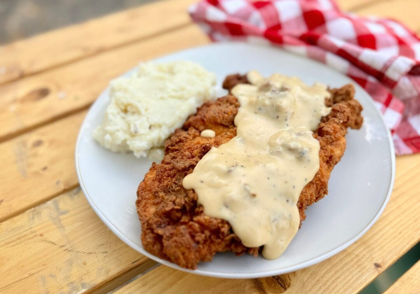 Cowboy Kent Rollins' Golden Crispy Chicken Fried Chicken with homemade gravy on a white plate, mashed potatoes on the side, with a red and white checked napkin in the background, on a picnic table.