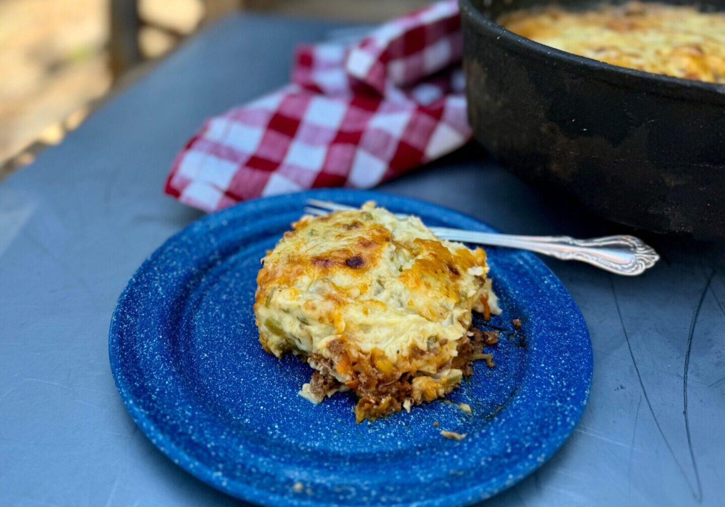 A blue enamel plate with cottage pie. In the background is a red checked cheesecloth and a Dutch oven. Cowboy Kent Rollins' Cottage or Shepherd's Pie