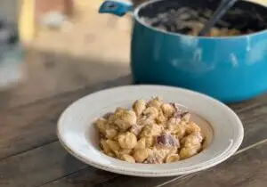 In the background, a blue enamel stock pot holds creamy, delicious smoked mac and cheese. In the foreground of the photo, a white plate holds smoked mac and cheese with smoked breakfast sausage. It is a miracle the pups didn't get to it before we got these photos taken.